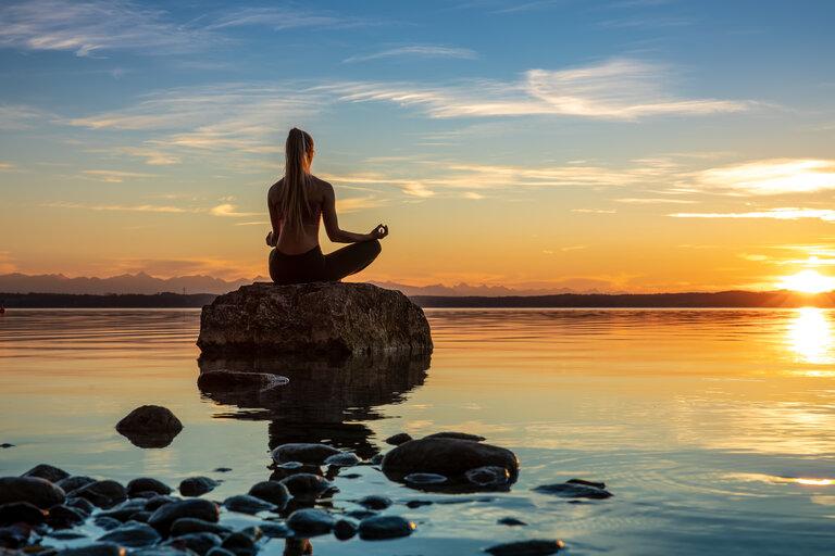 hübsche junge Frau macht Yoga auf einem Stein im Wasser, am See oder Meer bei Sonnenuntergang