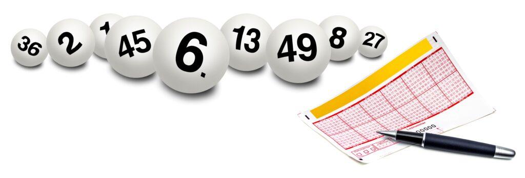 Weiße Lottokugeln mit schwarzen Zahlen in einer Reihe und ein ausgefüllter Lottoschein mit einem schwarzen Kugelschreiber daneben.