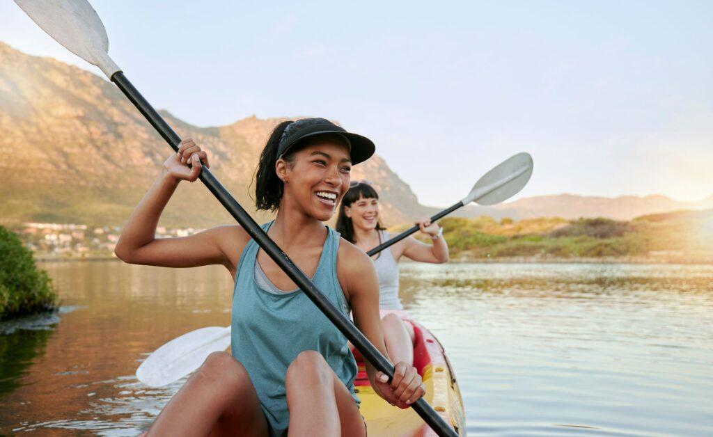 Zwei lächelnde Freunde Kajakfahren auf einem See zusammen während der Sommerpause. Lächelnde und glückliche spielerische Frauen, die sich draußen in der Natur mit Wasseraktivitäten verbinden. Spaß haben auf einem Kajak während der Wochenendfreizeit