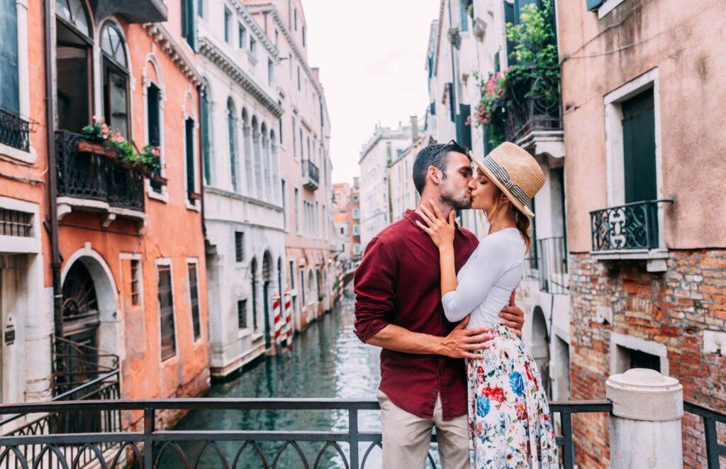 Ein verliebtes Paar küsst sich leidenschaftlich auf einer Brücke in Venedig mit historischen Gebäuden im Hintergrund, während sie exklusive Momente mit Amorette International genießen.