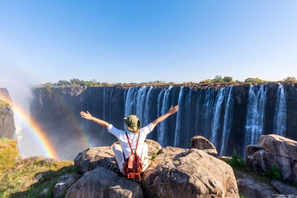 Person sitzt vor den beeindruckenden Victoriafällen mit Regenbogen – ein Highlight auf einer Reise durch Afrika, ergänzend zur Tansania Safari.