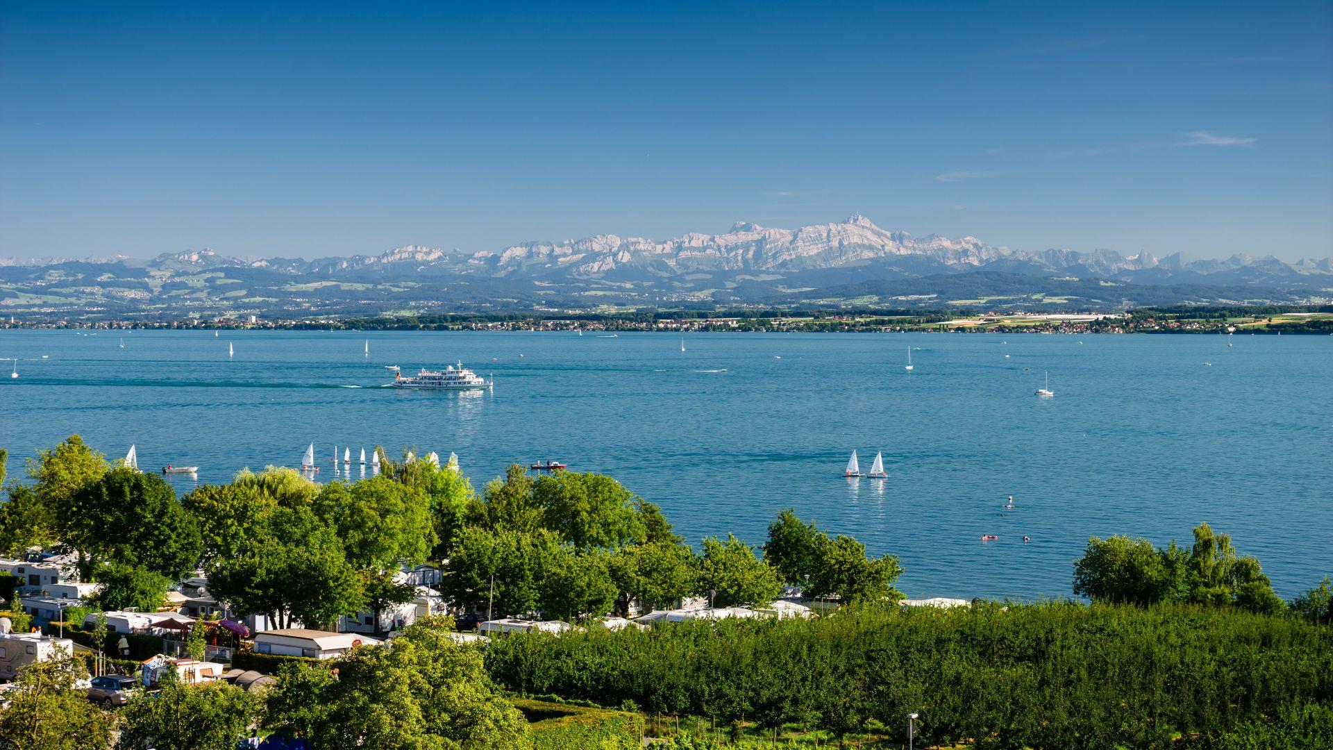 Panoramablick über den Bodensee mit Alpen im Hintergrund und Segelbooten auf dem Wasser