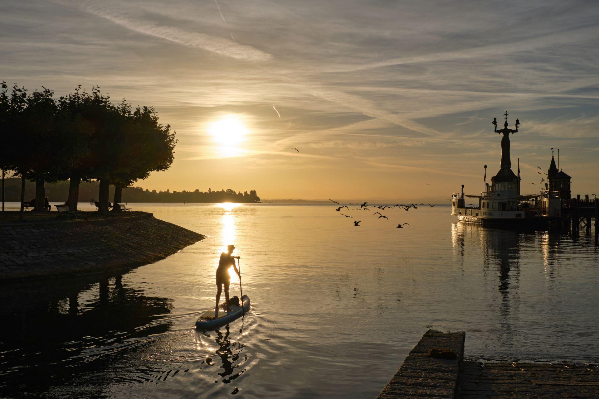 Person beim Stand-up-Paddling bei Sonnenaufgang am Bodensee mit Blick auf den Hafen von Konstanz